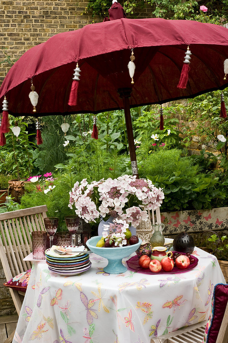 Gartentisch mit Hortensien, Obst, Tellerstapel und Gläsern unter Sonnenschirm