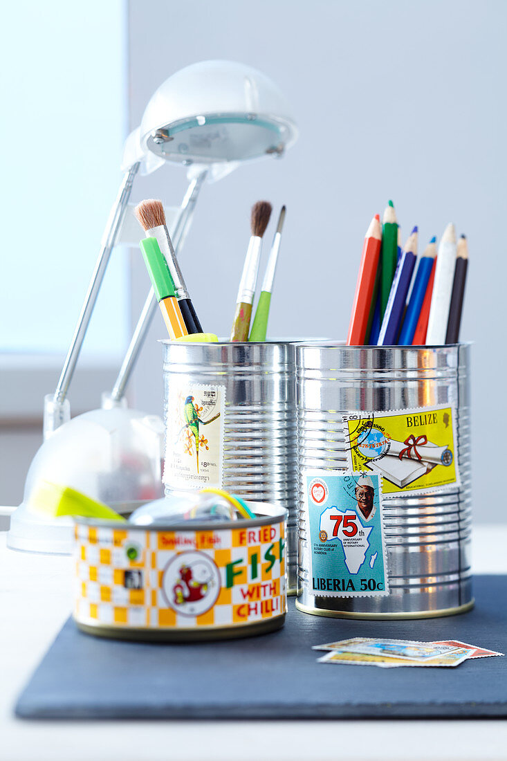 Coloured pencils and paintbrushes in decorated tin cans on a desk