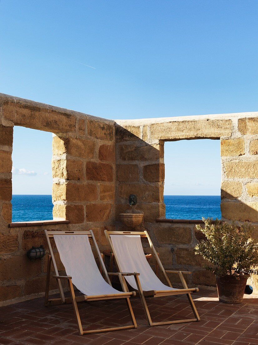 Wooden deckchairs with pale canvas seats on Mediterranean terrace surrounded by high stone wall with openings showing sea view
