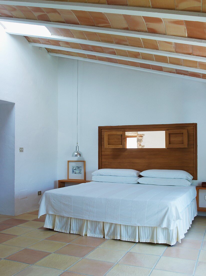 Double bed with valance and white bedspread in simple bedroom with ceiling of roof tiles