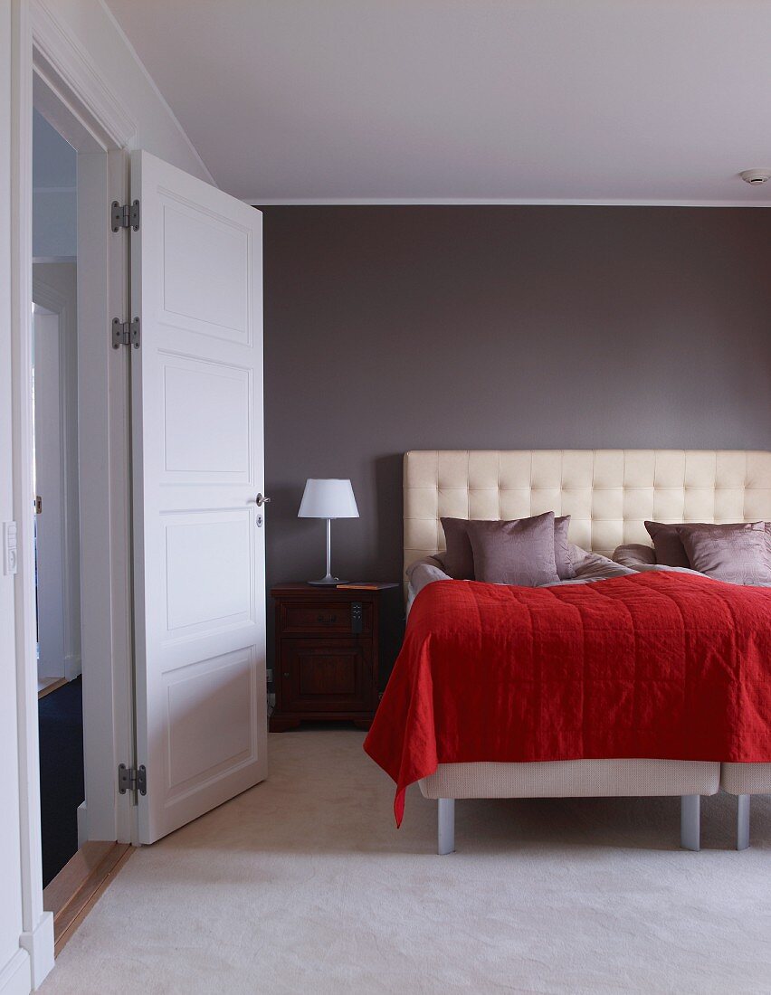 Bett mit gepolstertem Kopfteil und roter Tagesdecke vor grauer Wand in modernem Schlafraum mit offen stehender Tür