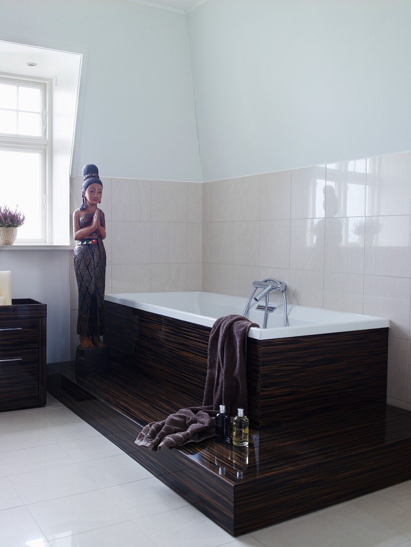 Statue next to bathtub on platform clad in exotic wood against white-tiled wall