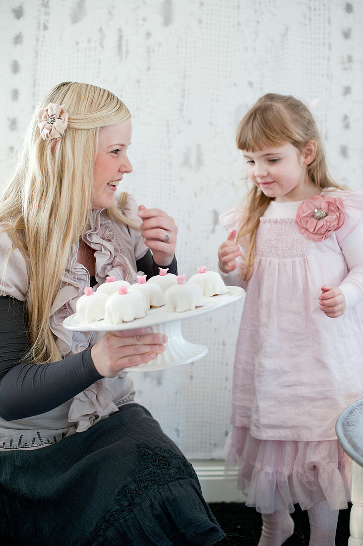A mother dressed for a celebration, with her young daughter, holding a cake stand of mini birthday cakes in her hand