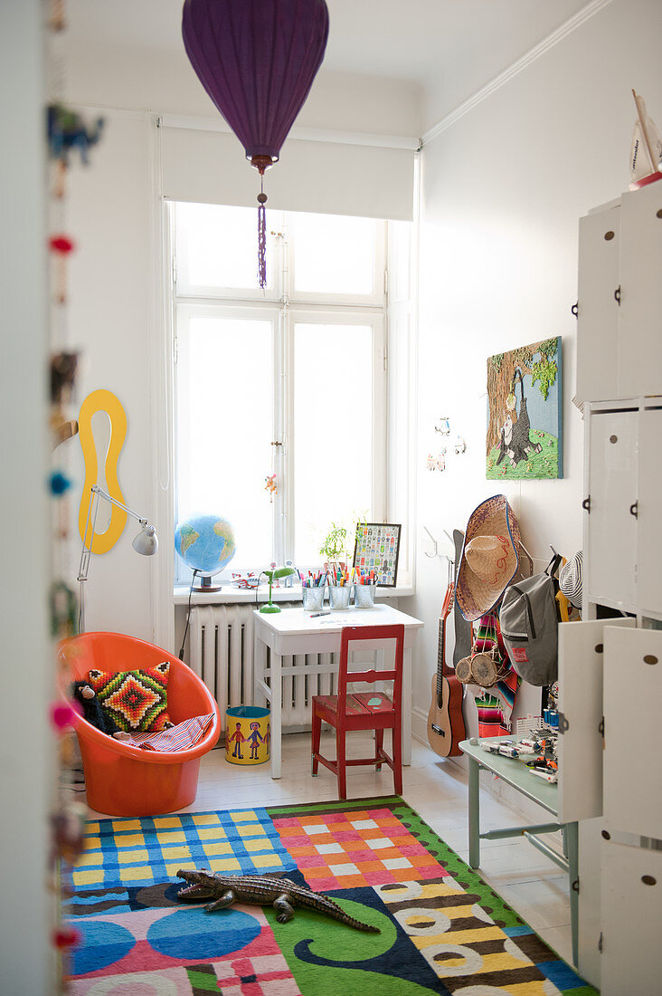 Helles Kinderzimmer mit buntem Teppich und roter Stuhl vor kleinem Tisch am Fenster