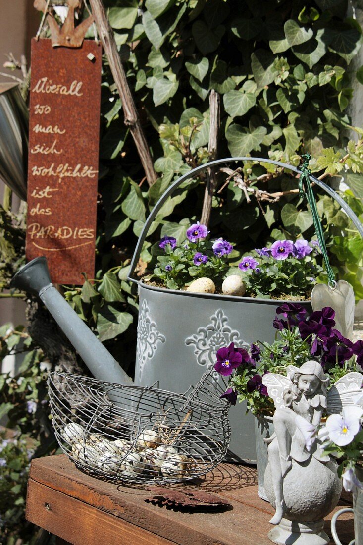 Zinkgiesskanne und Elfe mit Hornveilchen und Korb mit Wachteleiern auf einem Gartentisch
