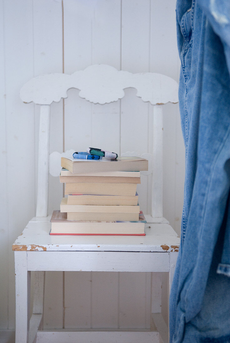 Bücherstapel auf weiss lackiertem Küchenstuhl mit Schnitzereien auf Rückenlehne vor weisser Holzverkleidung