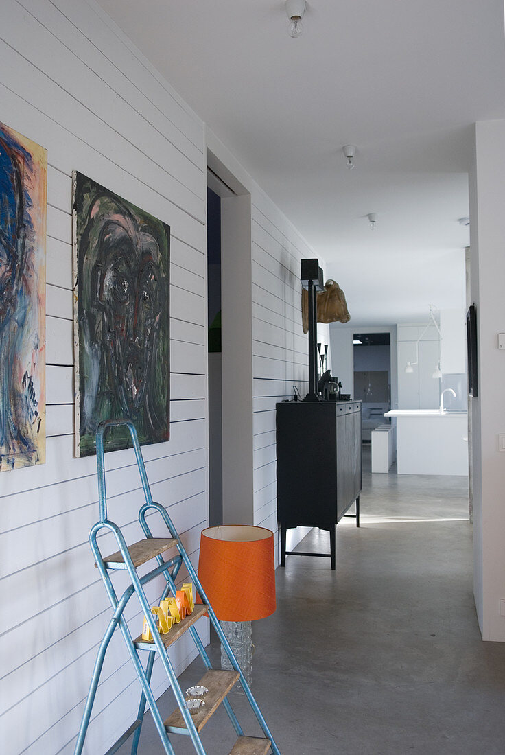 Trittleiter vor modernen Gemälden an holzverkleideter Wand und 70er Jahre Lampe mit orangefarbenem Schirm