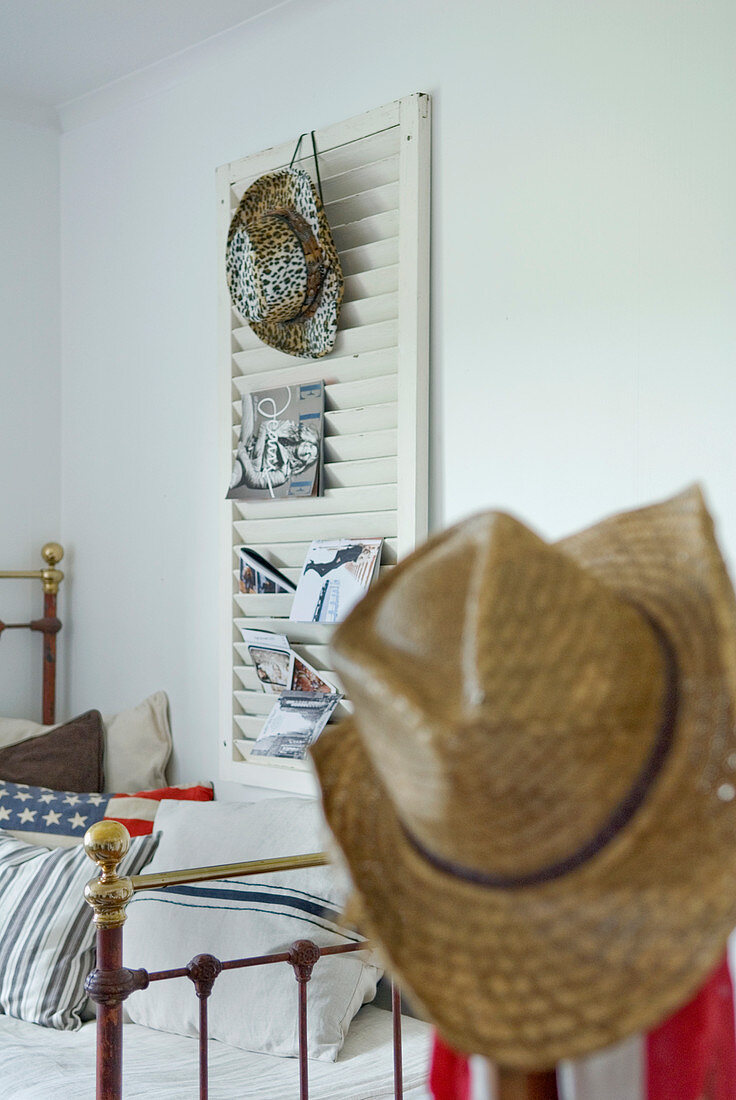 Schlafzimmer mit Strohhut, Vintage Metallbettgestell und Cowboyhut mit Zeitschriften an Deko-Jalousiefensterladen an weißer Wand