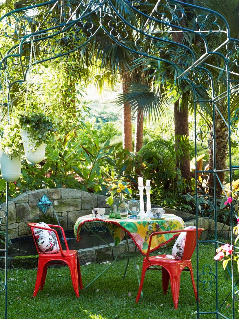 Gartentisch mit roten Stühlen unter Rosenbogen aus Metall, im Hintergrund Steinbrunnen