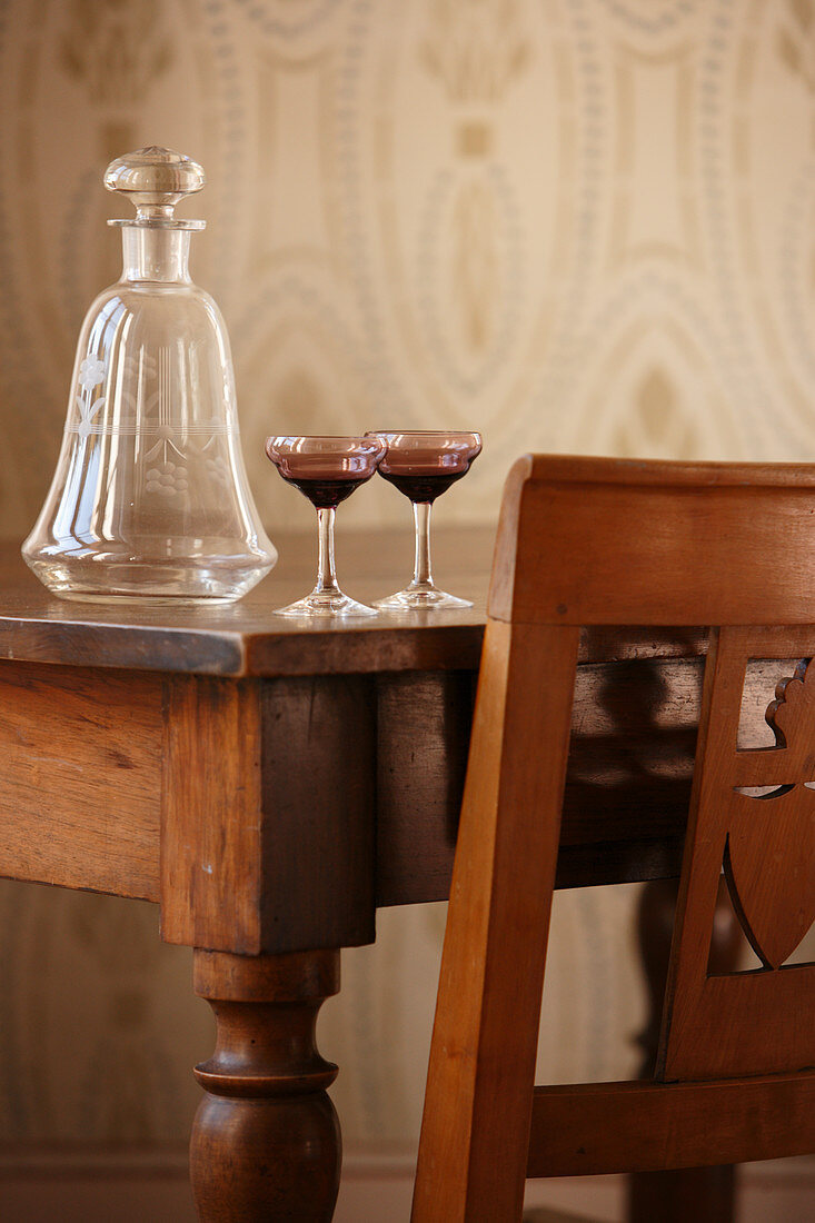 Antike Glaskaraffe und Gläser auf dem Holztisch und teilweise sichtbarer Stuhl mit geschnitzter Rückenlehne