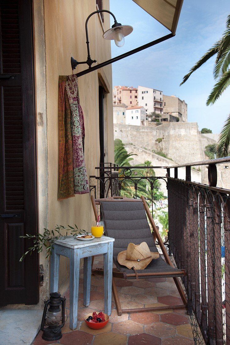 Schlichtes hellblaues Beistelltischchen neben grauem Holzliegestuhl auf kleinem Balkon eines mediterranen Wohnhauses mit Blick auf die Stadt am Hang gelegen.
