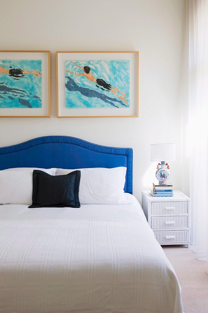 White bedspread on double bed with blue upholstered headboard below modern artworks on wall
