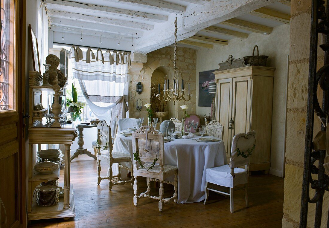 Mit einer Sammlung antiker Stühle, Kronleuchter und bodenlangem Tischtuch festlich gedeckte Tafel in einfach restauriertem Landhaus
