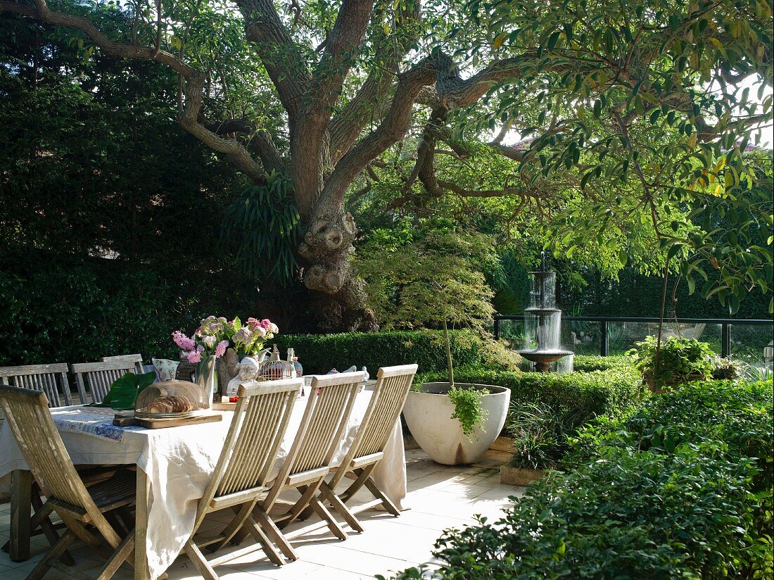 Gedeckter Tisch unter einem Baum im Garten, im Hintergrund Springbrunnen