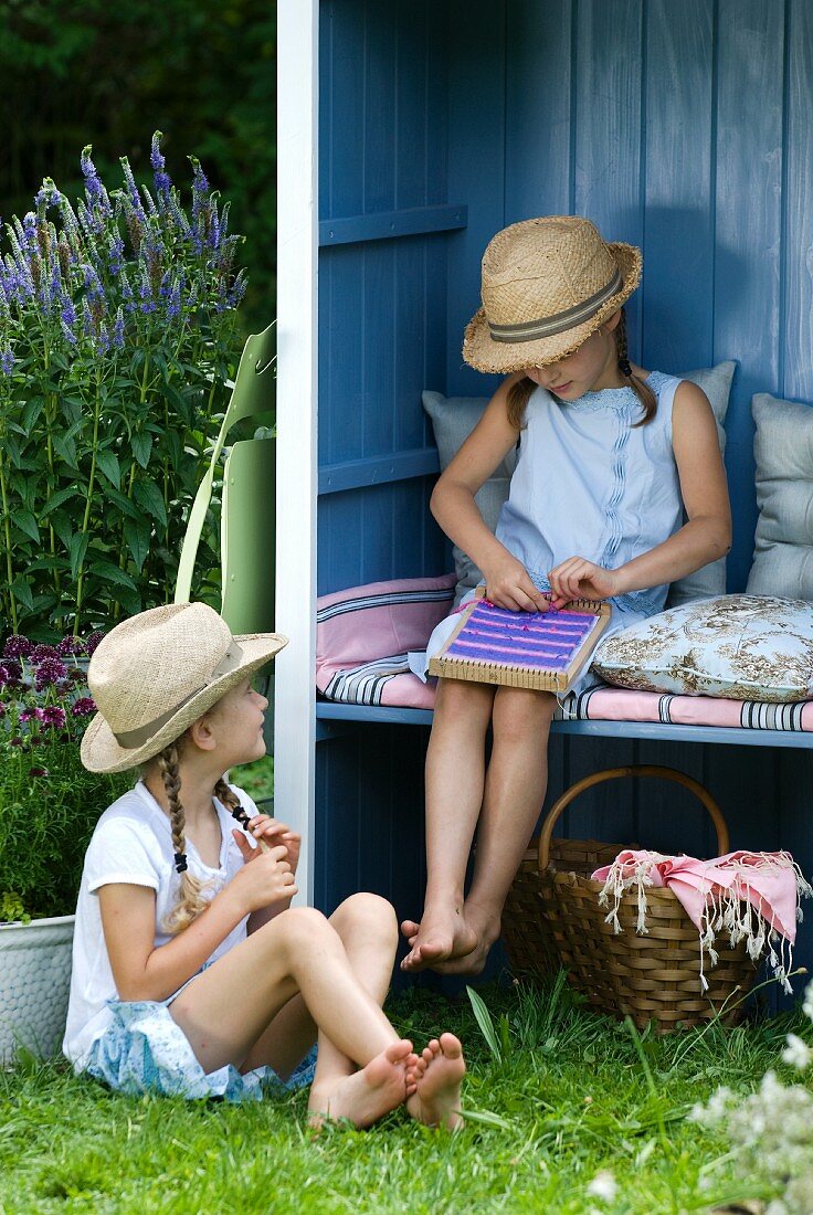 Girl with weaving frame sitting in open-fronted garden shed behind second girl sitting on lawn
