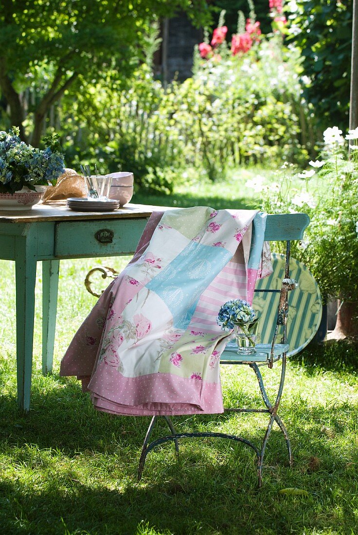 Patchwork blanket with delicate patterns draped over garden chair in front of old wooden table in summer garden