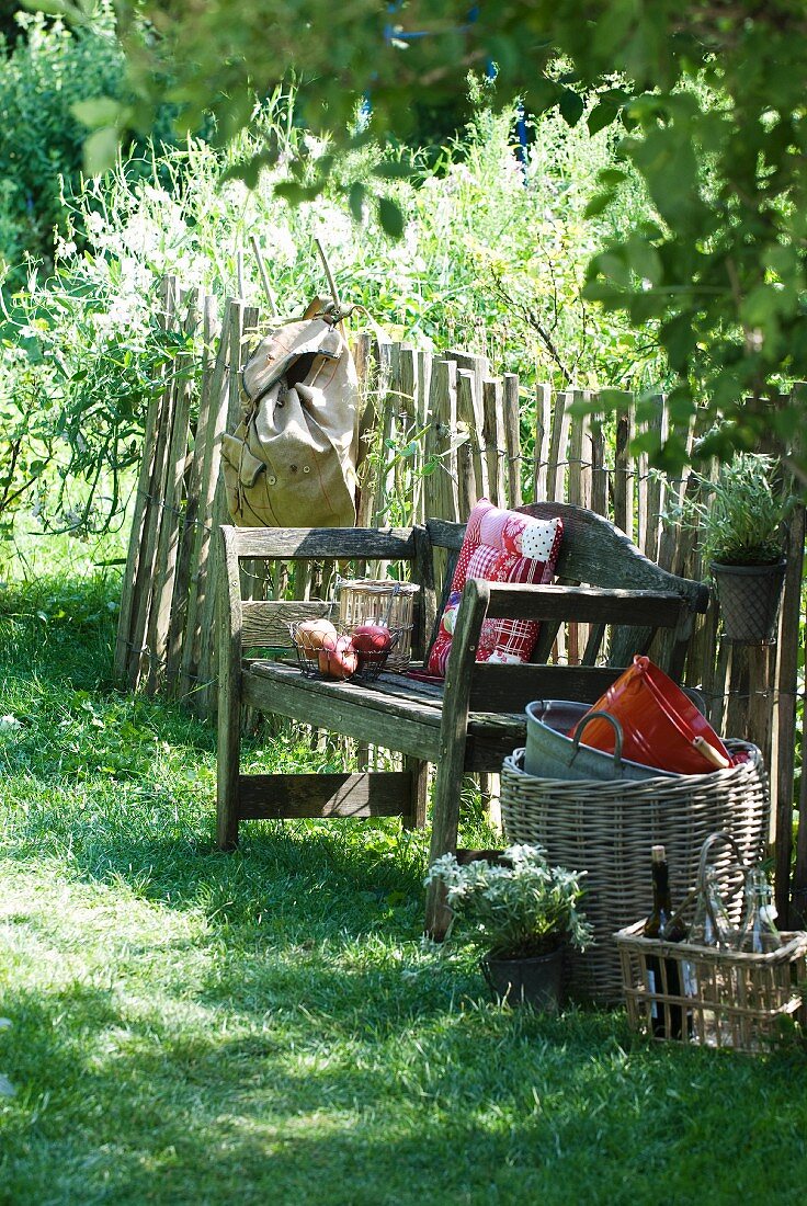 Rustic seating area in garden: old wooden bench with scatter cushions, backpack, wicker basket, basket of bottles