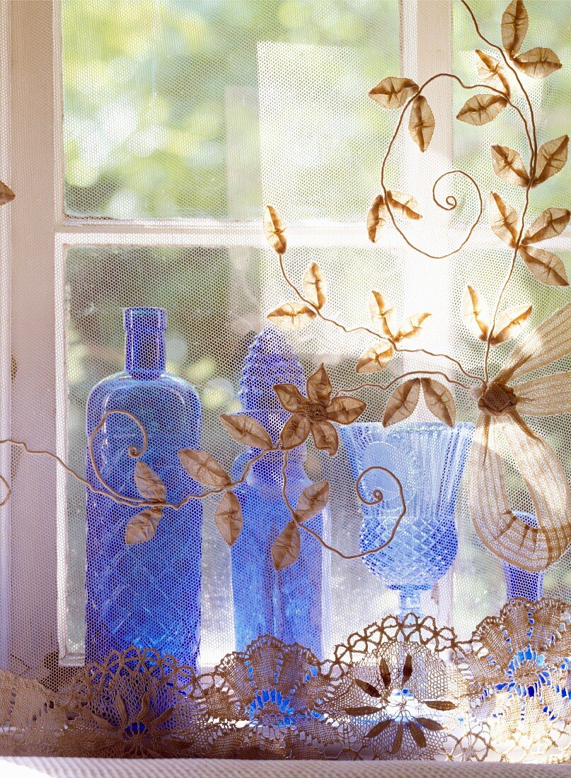 Blaue Glasvasen hinter transparentem Tüllvorhang mit floralen Stickereien