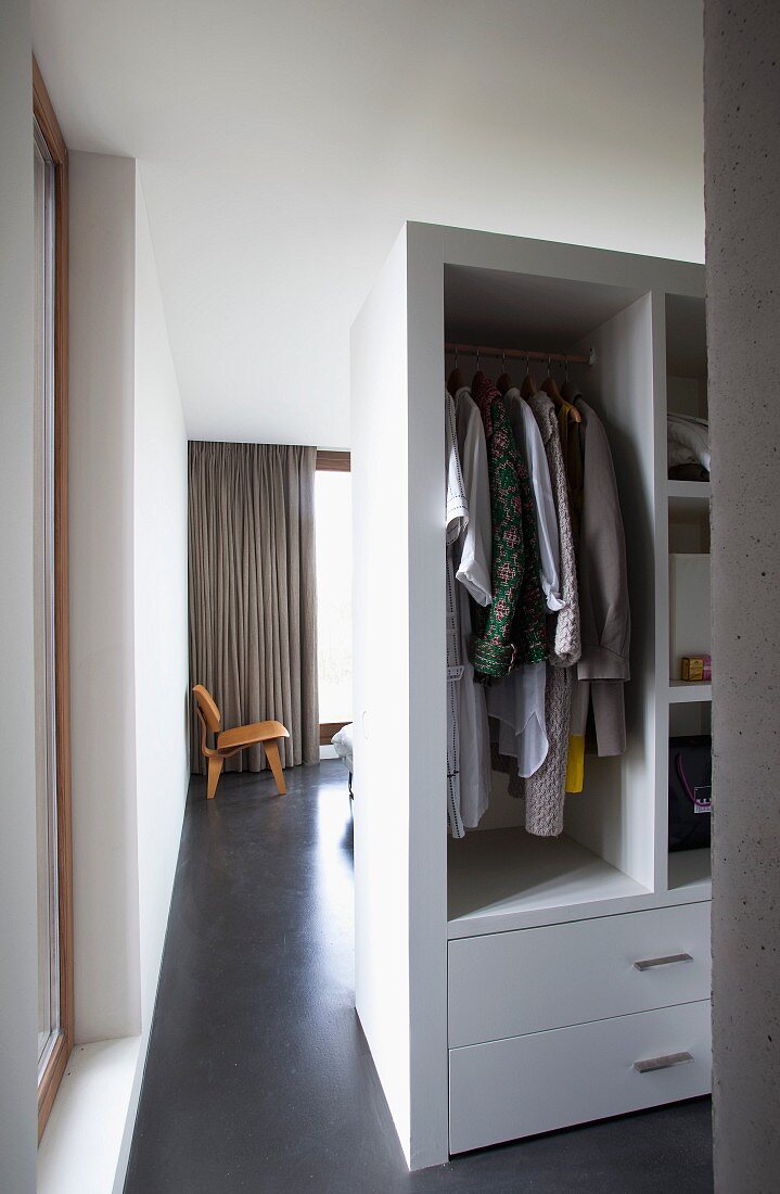 Blick vorbei am Raumteiler-Kleiderschrank mit offenen Fächern in das Wohnzimmer mit Designerstuhl auf poliertem Betonboden