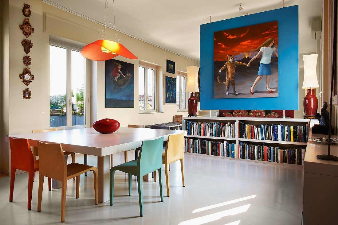Moderner Esstisch und farbige Stühle vor blauem Raumteiler mit grossformatigem Bild über Lowboard mit Büchern in offenem Wohnraum