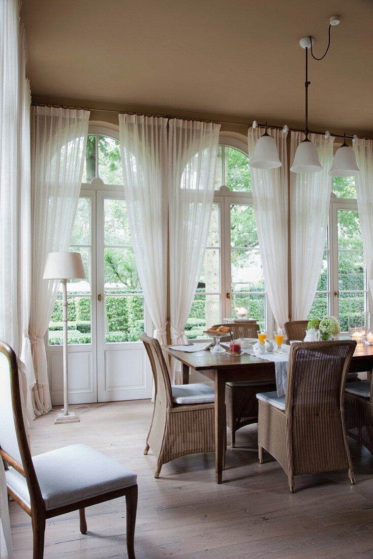 Wicker chairs around breakfast table in front of arched French windows in dining room with traditional ambiance