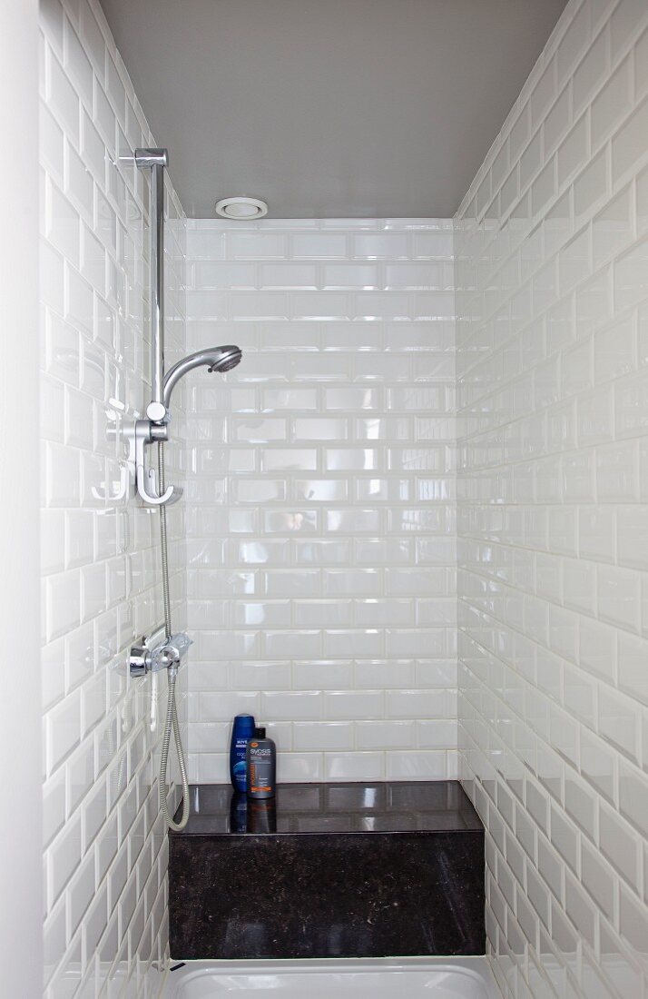 Narrow shower with white wall tiles and toiletries on black stone bench