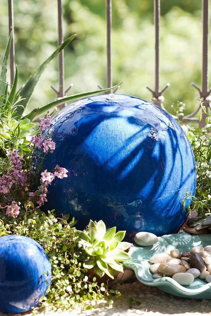 Blue-glazed, ceramic, spherical water feature amongst ground cover plants and decorative pebbles against balcony railings