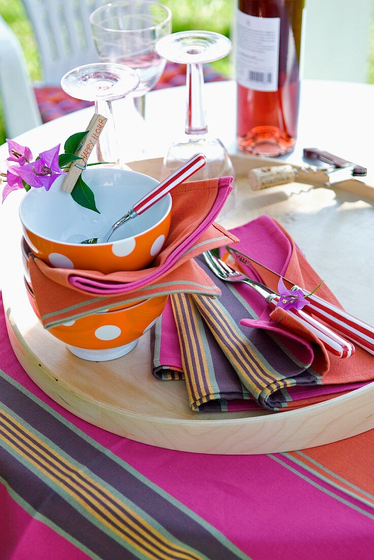 Spotty bowls, colourful fabric place mats and striped cutlery on wooden tray in garden