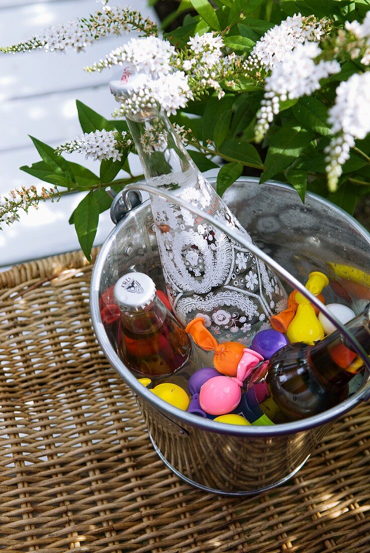 Dekorative Getränkeflaschen zum Kühlen zwischen bunten Eiswürfel-Ballons in Metalleimer; weiße Blüten im Hintergrund