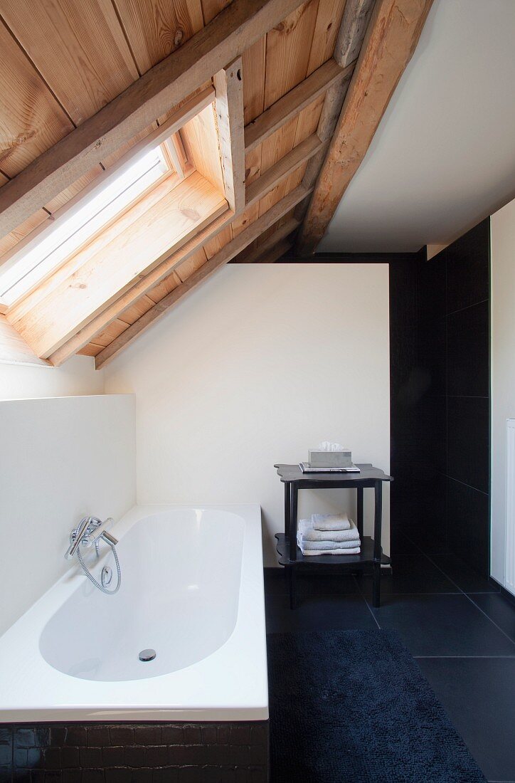 Designer bathroom in renovated attic - black tiled floor and bathmat next to bathtub below skylight