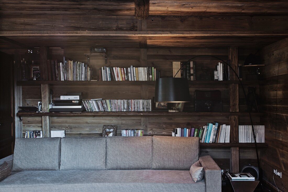 Holzverkleidete Wand mit Bücherregal; im Vordergrund eine graue Couch und eine schwarze Bogenlampe