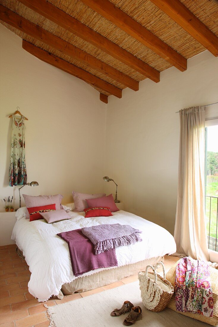 Exposed roof structure with straw mats in pleasant, bright bedroom; red and purple cushions and blankets on French bed