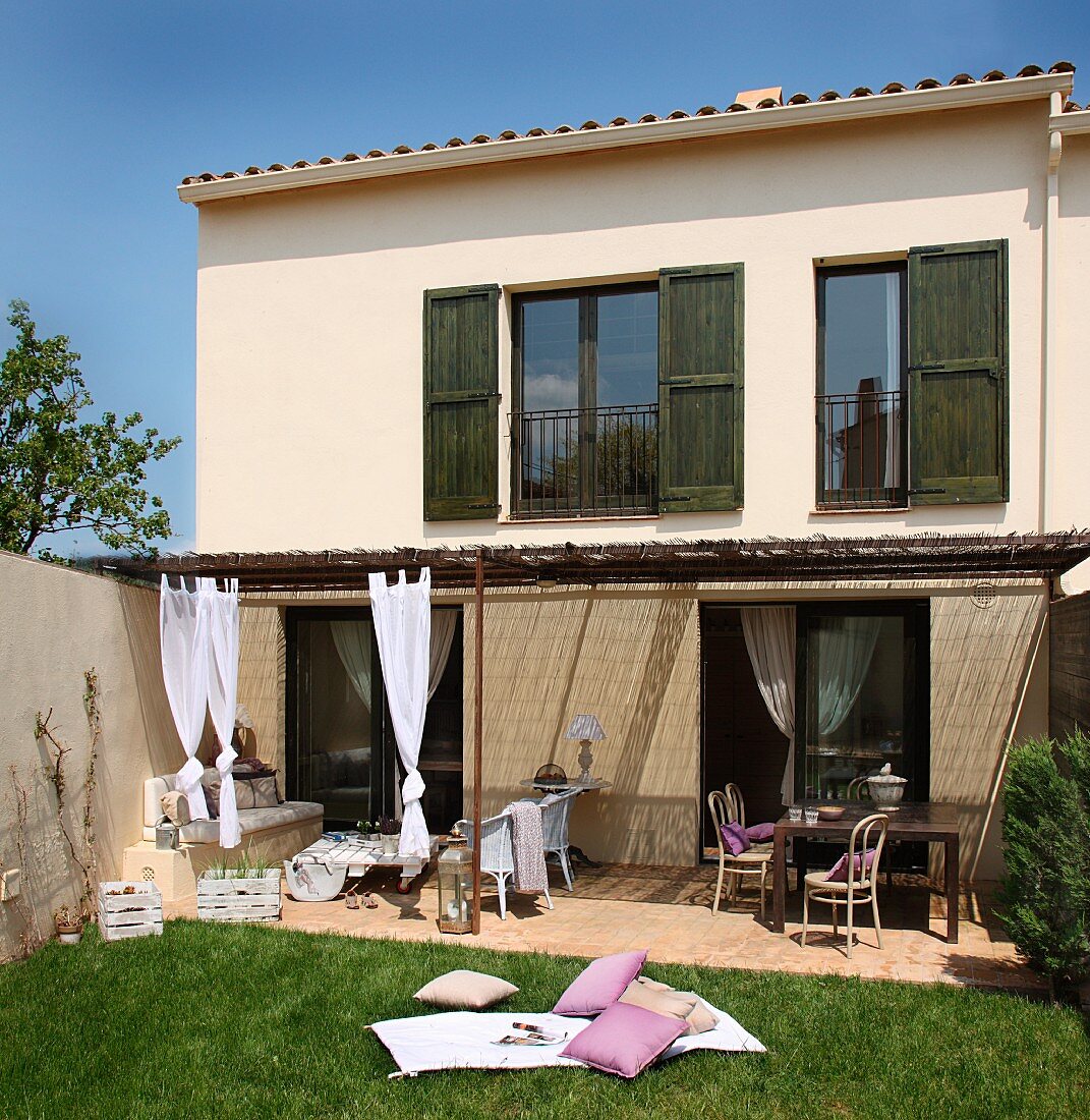 Simple Spanish holiday home with dark green shutters and straw-thatched pergola with draped curtains above furnished terrace