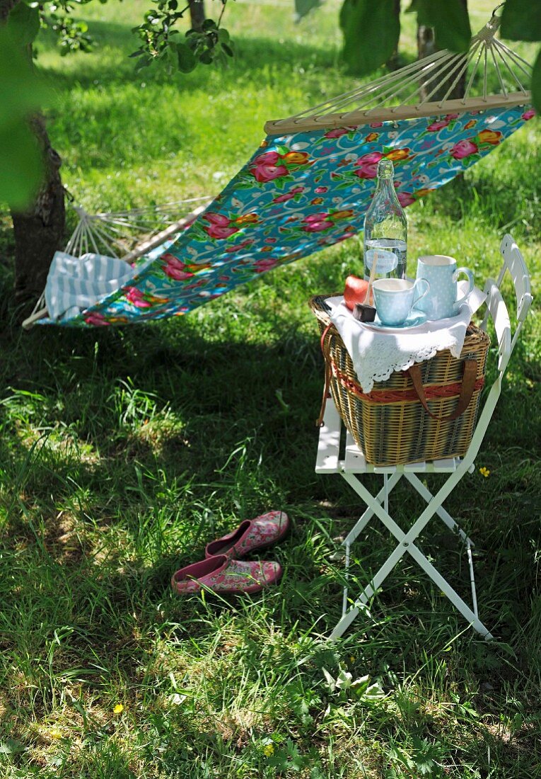 Picknickkorb auf Stuhl und zwischen Bäume gespannte Hängematte