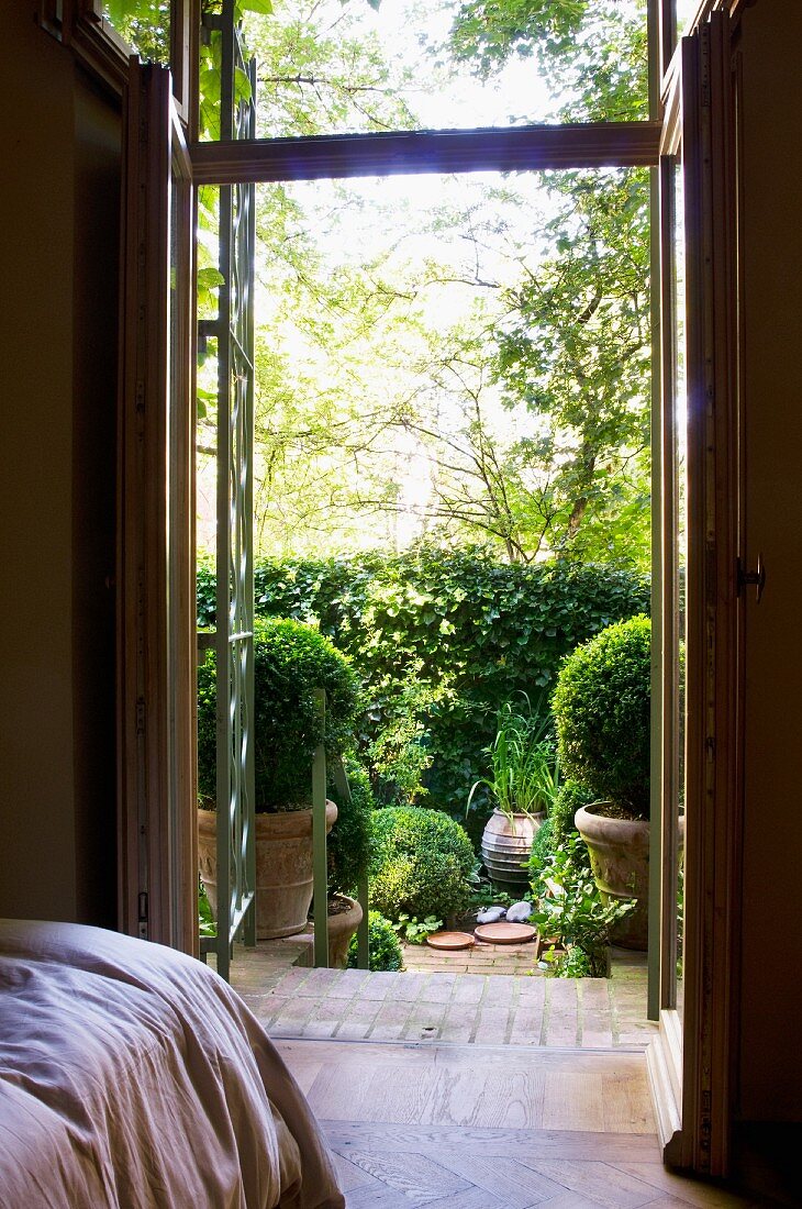 Blick vom Schlafzimmer durch offene Terrassentür auf sonnenbeschienene Terrasse mit Buchsbaumkugeln in Töpfen