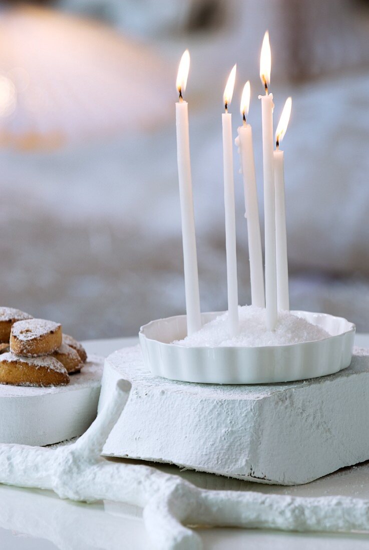 Schale mit aufgehäuftem Salz und hineingesteckten, brennenden Kerzen auf weiss gestrichenem Holzstück neben Süssigkeiten