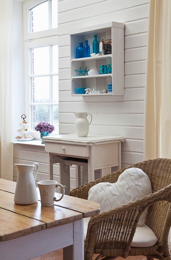 Blick über Küchentisch und Rattanstuhl auf Wandtisch und Regalschränkchen an weisser Holzwand