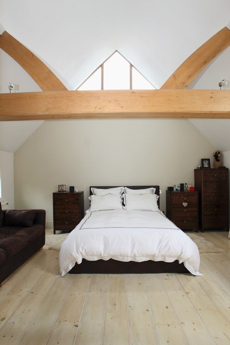 Weiß bezogenes Doppelbett und braune Holzkommoden in ausgebautem Dachraum mit offener Holzbalkenkonstruktion