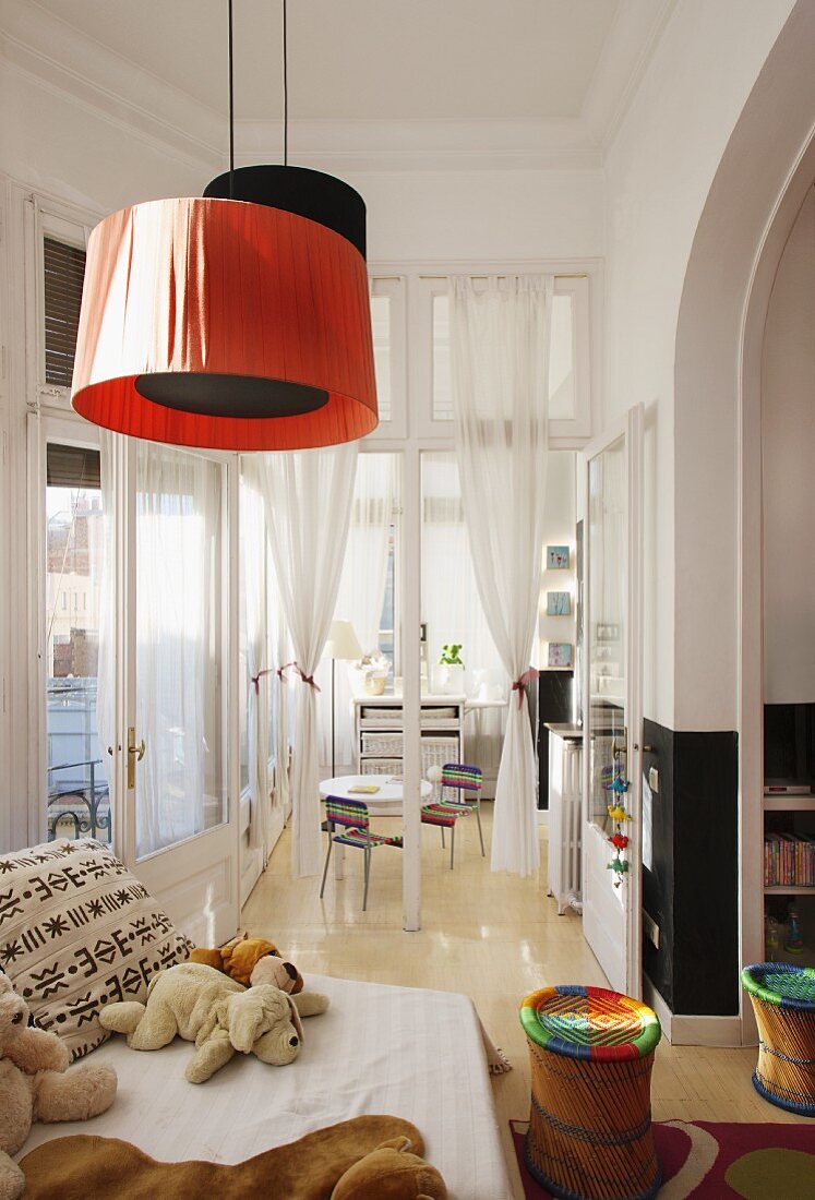 Soft toys on sofa, colourful stools and red and black pendant lamps in child's bedroom with open glass door leading to playroom in background