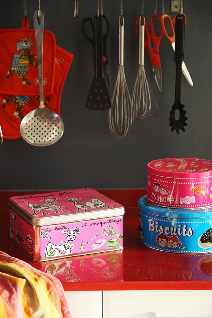 Colourful metal tins on red worksurface below kitchen utensils hanging against grey wall