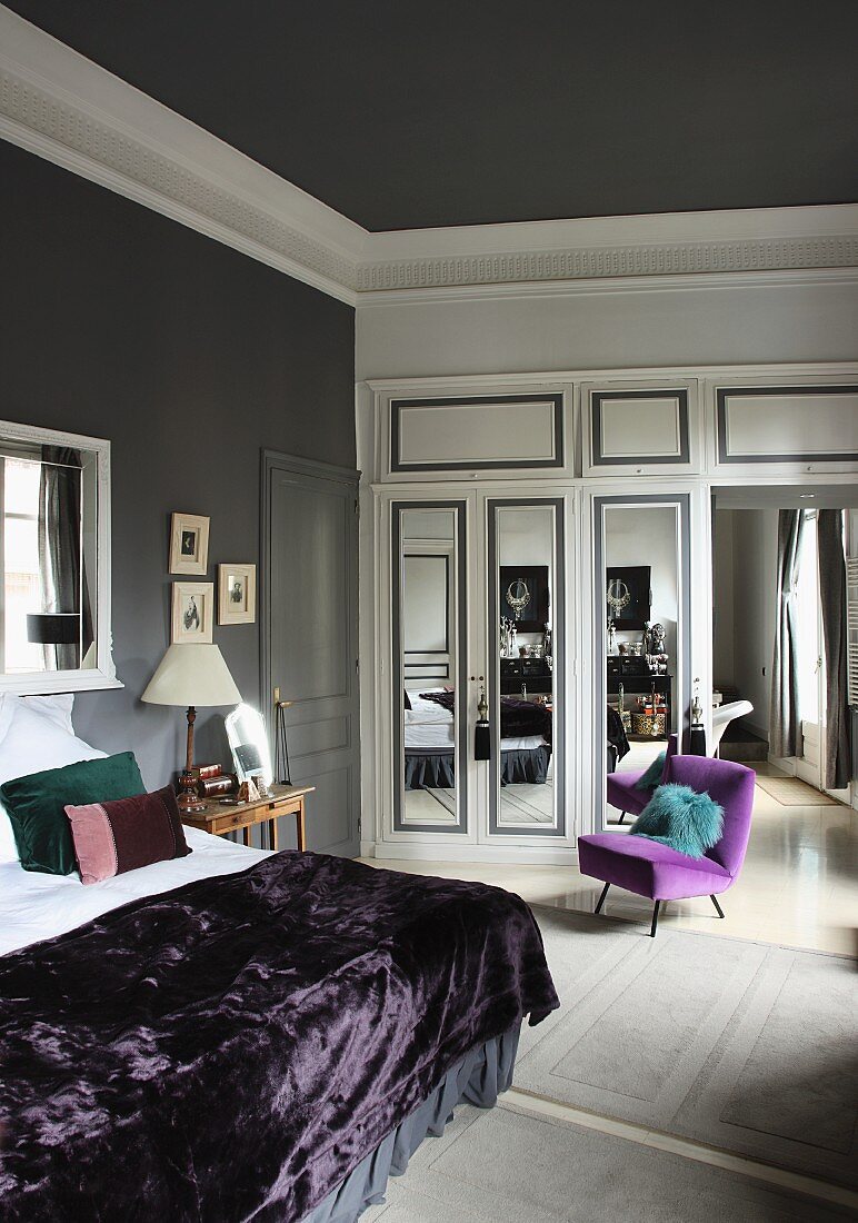 Doppelbett mit lila Tagesdecke, lila Polsterstuhl und weisser Einbauschrank mit Spiegeltüren in einem Schlafzimmer mit grauen Wänden