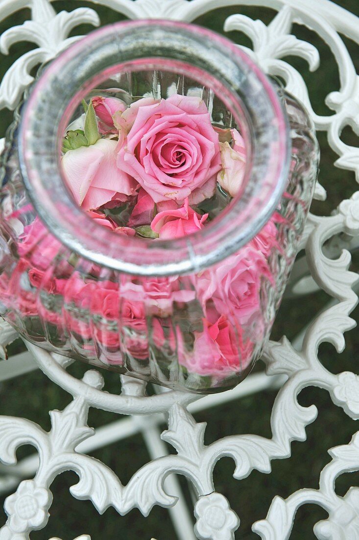 Glasbehälter mit Rosenblüten auf Ablage aus floralem, schmiedeeiesernem Gitter