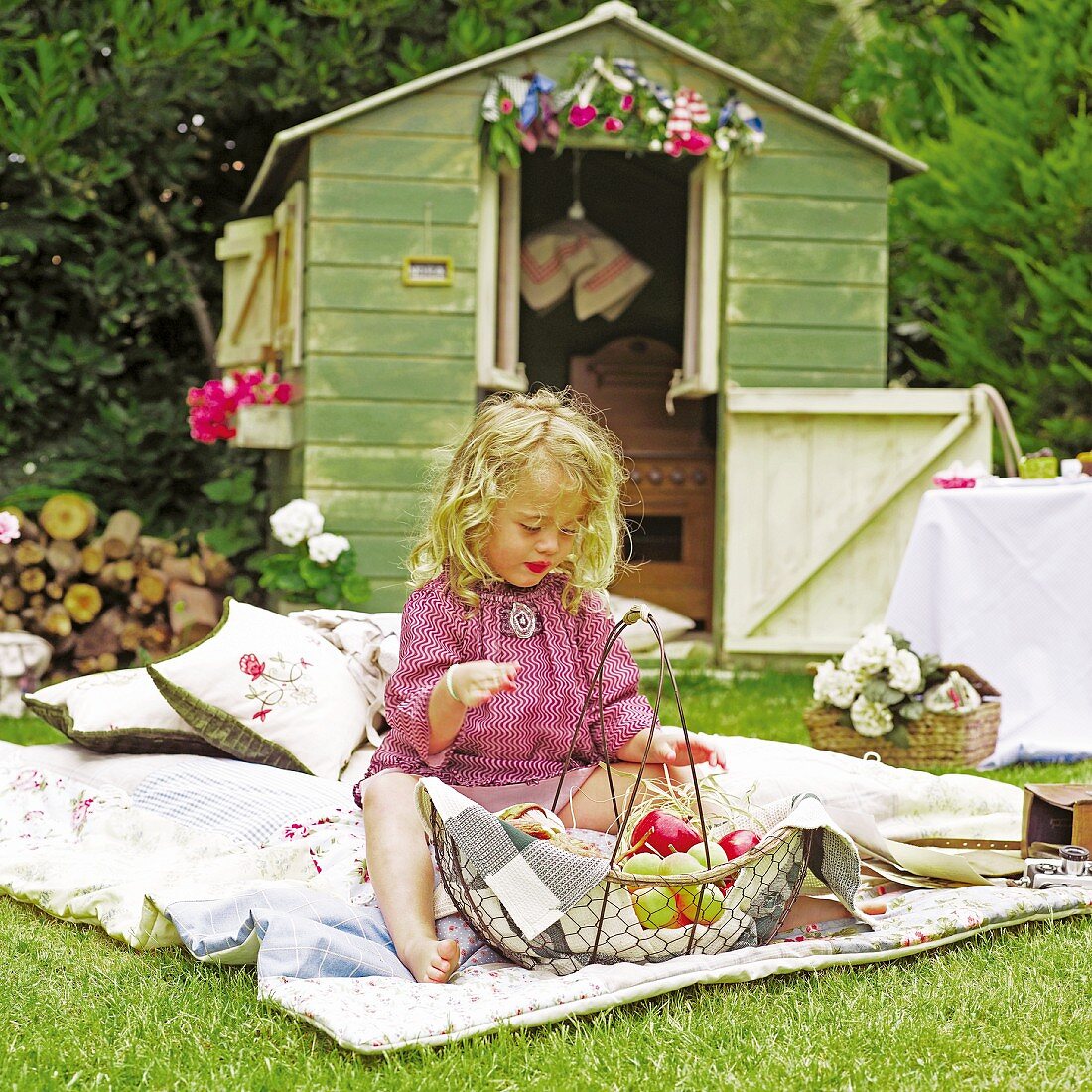 Kleines, blondes Mädchen auf Patchworkdecke mit obstgefülltem Drahtkorb; blumengeschmücktes Gartenhaus im Hintergrund