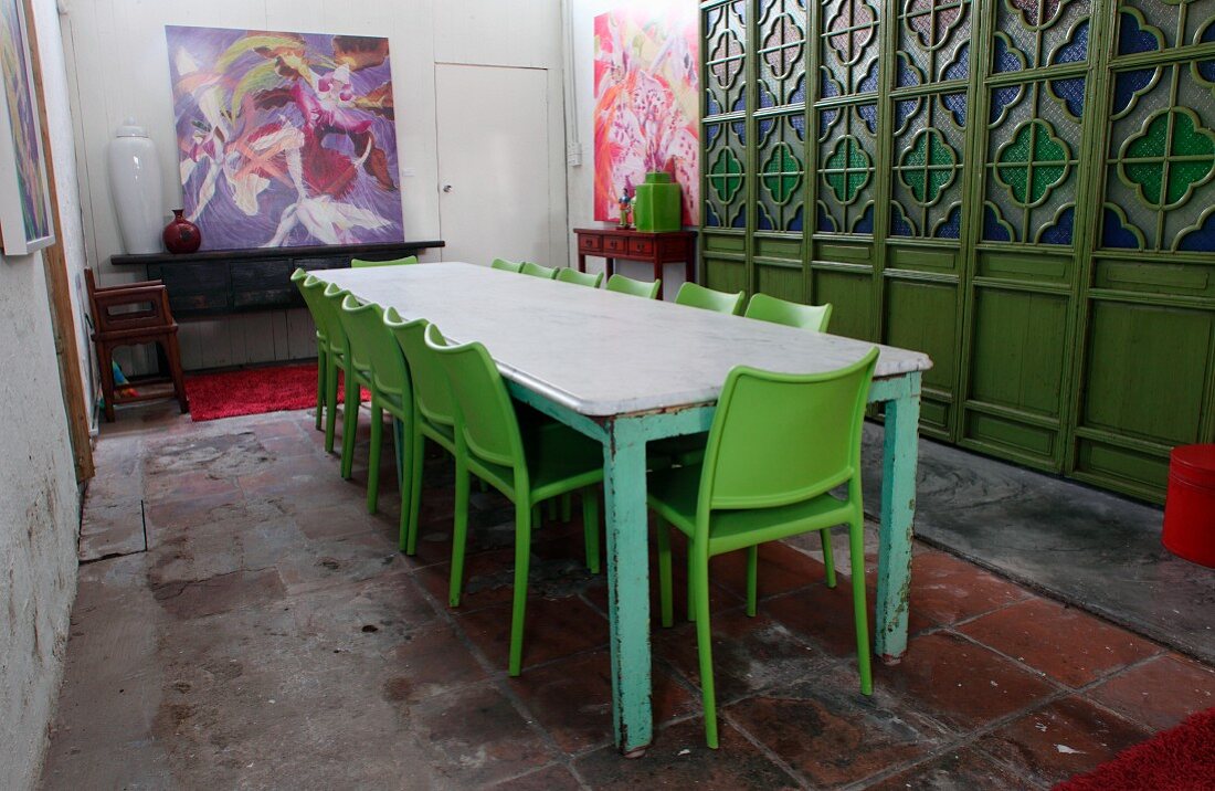 Long wooden table with peeling paint and green plastic chairs in front of wooden partition with stained glass windows in simple dining room