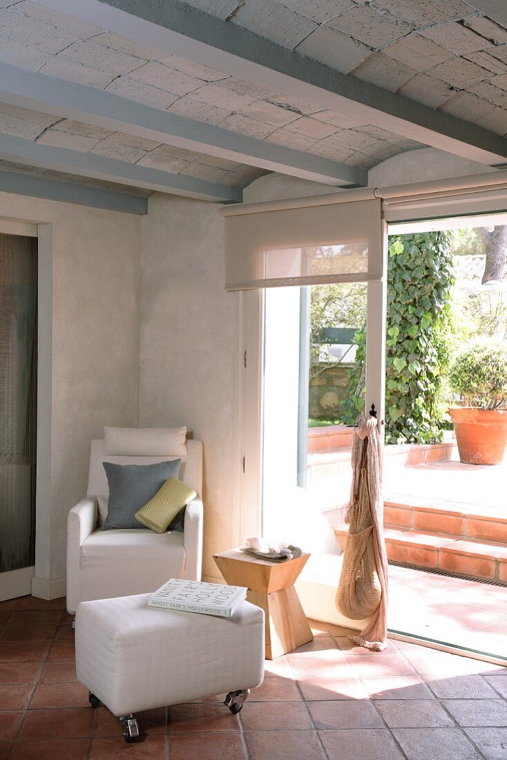 Heller Sessel und Polsterhocker mit Rollen vor offener Terrassentür in Wohnraum mit mediterranen Terrakottafliesen und unverputzter Rippendecke