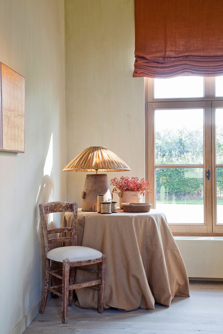 Runder Tisch mit bodenlanger Tischdecke und Tischlampe neben Fenster in Zimmerecke eines schlichten Wohnraumes