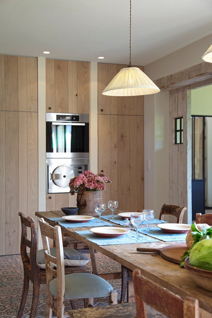 Gedecke auf Holztisch mit schlichter Hängelampe vor modernem Einbauschrank und integrierten Küchengeräten