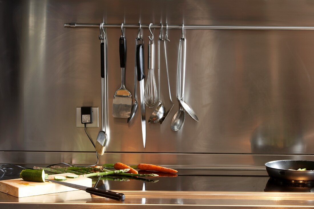 Küchenutensilien hängen vor Edelstahl Spritzschutzwand und Gemüse auf der Arbeitsplatte