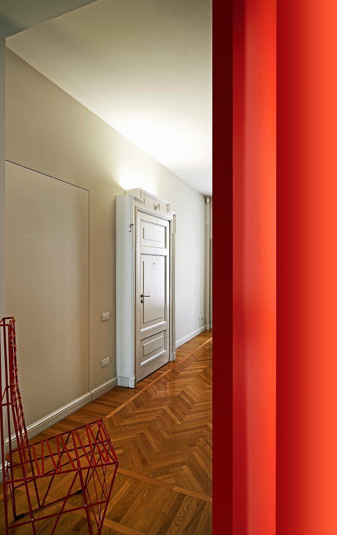 View of red wire-framed chair and herringbone parquet in grand hallway though doorway next to red wall