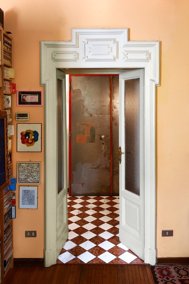 Offene Zimmertür mit geschnitztem weißem Holzrahmen in apricotfarbener Wand und Blick in Vorraum auf Schachbrettmusterboden